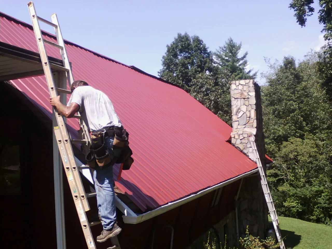 Roofer on a ladder beside a red metal roof and stone chimney — Soffit & Fascia Repair by Roofing Brothers in Crystal Beach